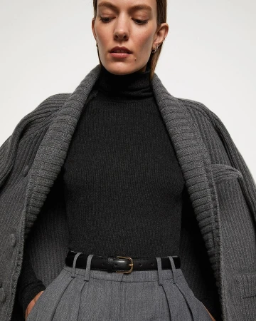 A sophisticated fashion image featuring an adult woman in a dark turtleneck, grey trousers, and a stylish grey cardigan against a plain background.