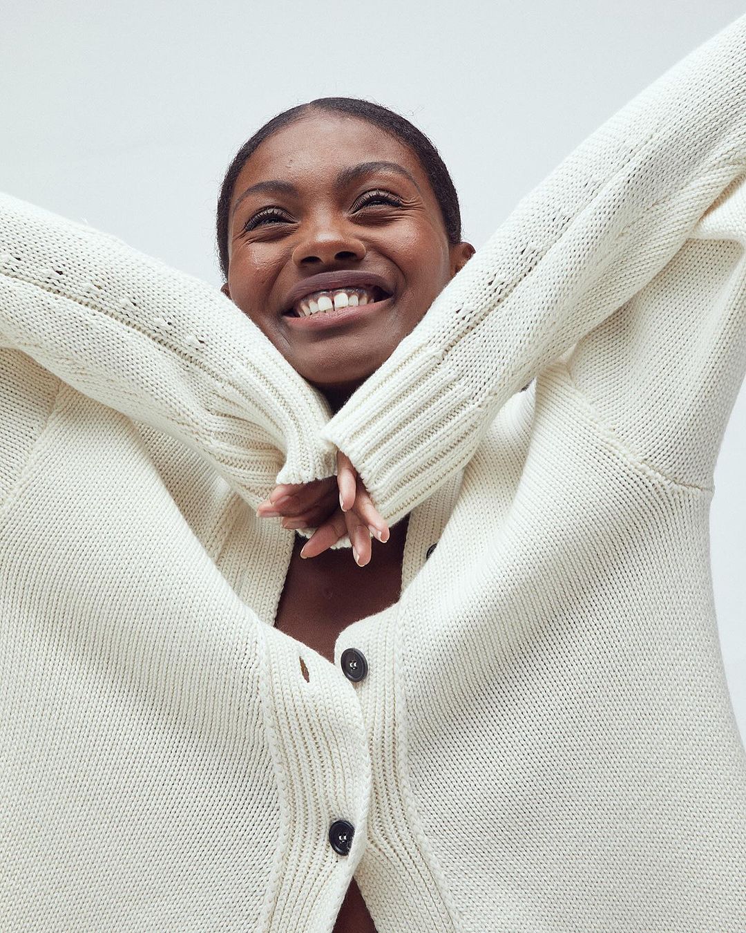 Smiling Black woman in a cream-colored knit button-down cardigan with arms raised in a low-angle portrait against a white backdrop.