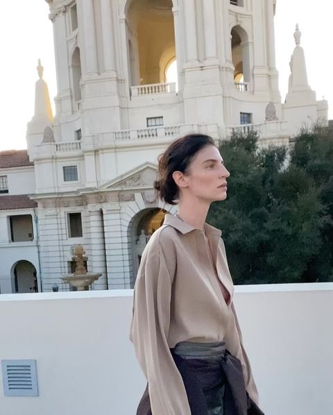 Woman poses in front of a large building with classical architecture on a sunny day, wearing a beige shirt and a dark wrap.