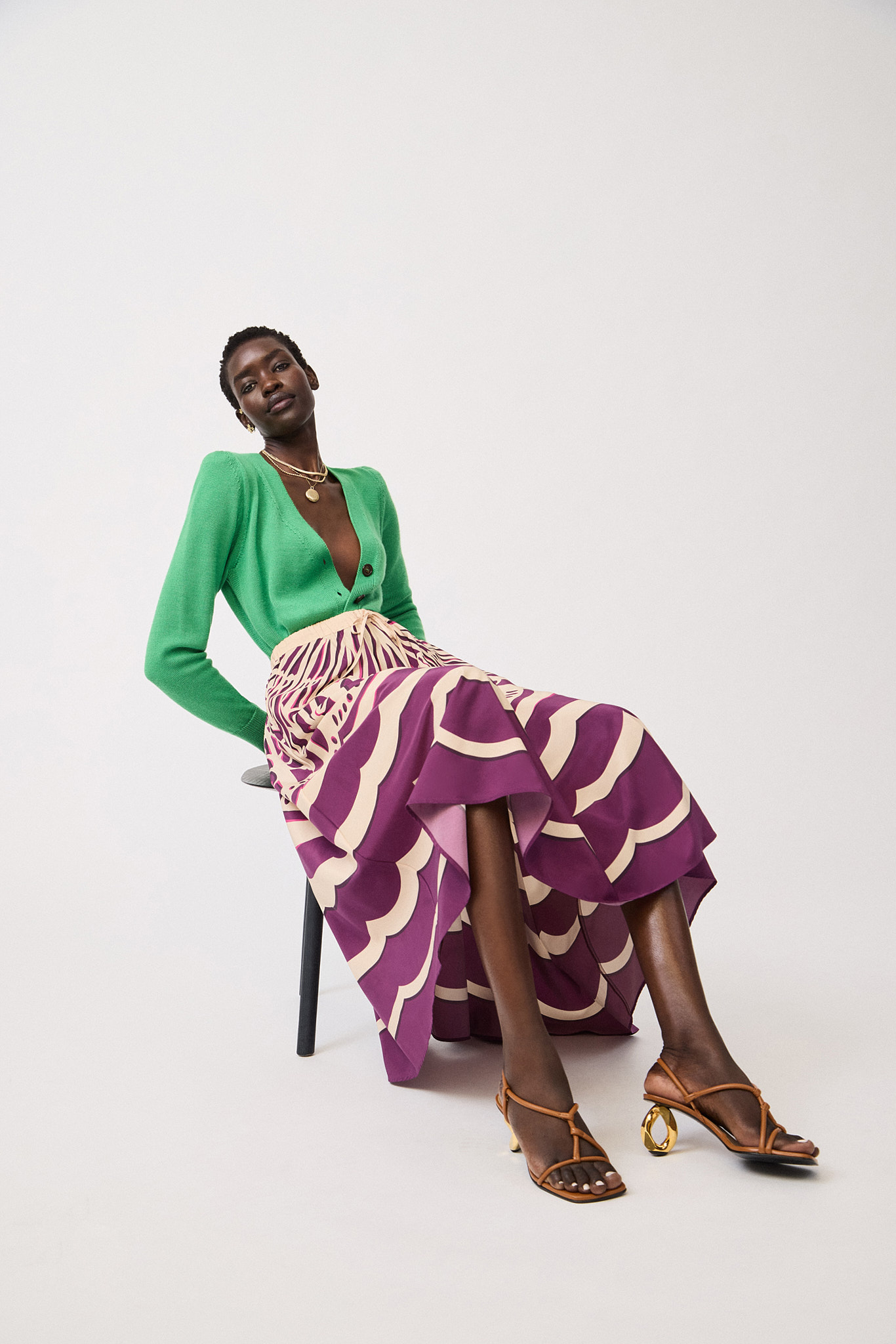 A Black female fashion model poses while seated, wearing a green cardigan, printed midi skirt, and strappy heeled sandals against white.