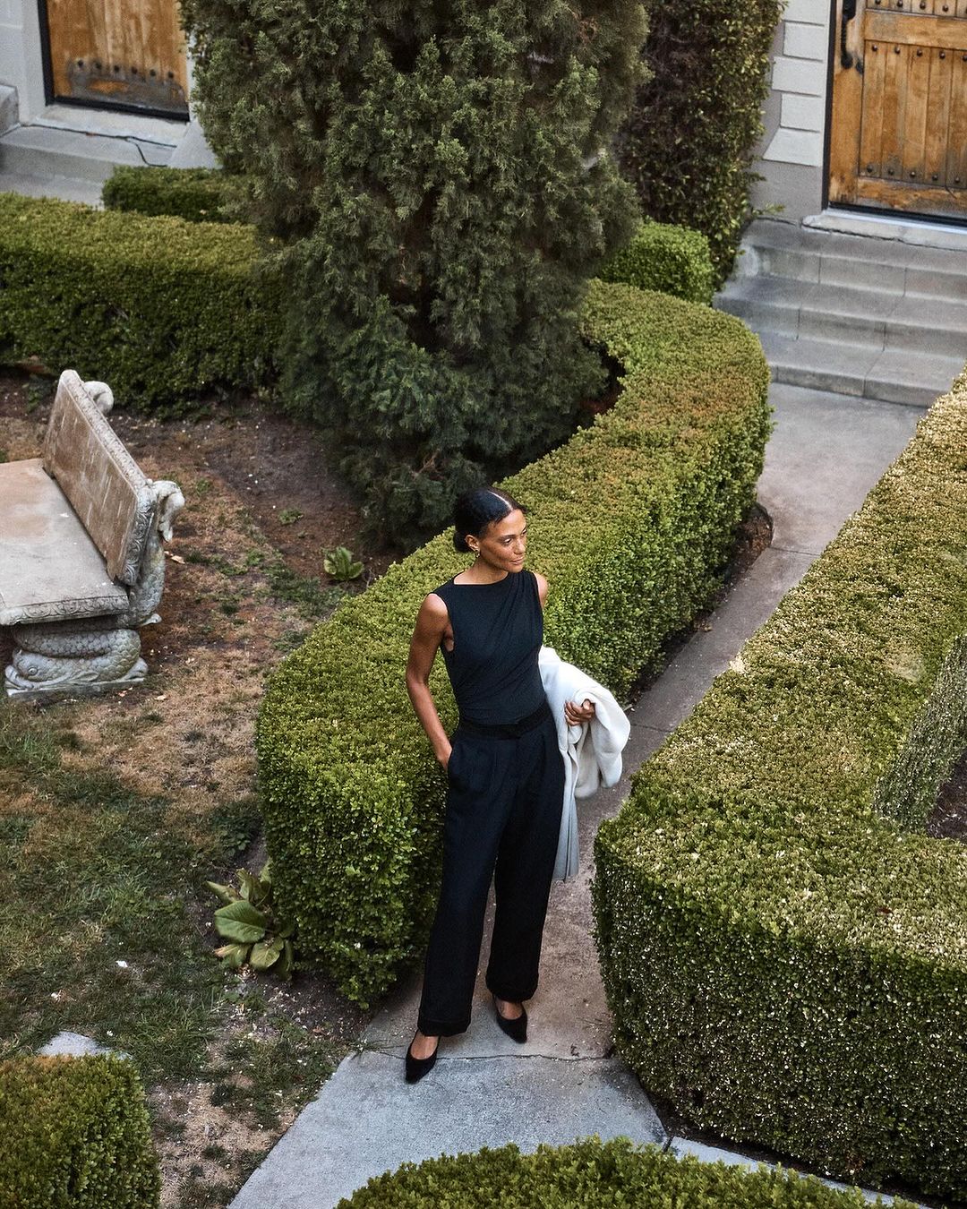 Elegant adult woman in a sleeveless black outfit poses amid sculpted green hedges in an outdoor garden on a concrete path.