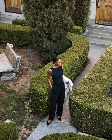 Elegant adult woman in a sleeveless black outfit poses amid sculpted green hedges in an outdoor garden on a concrete path.