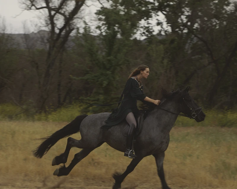 A woman rides a grey horse at a fast gallop across a grassy field with trees in the background on an overcast day.