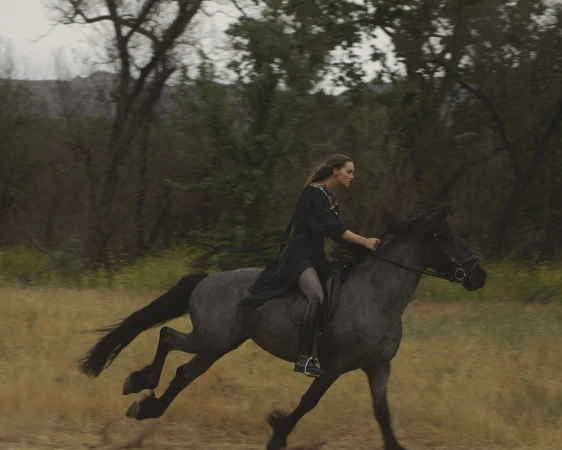 A woman rides a grey horse at a fast gallop across a grassy field with trees in the background on an overcast day.
