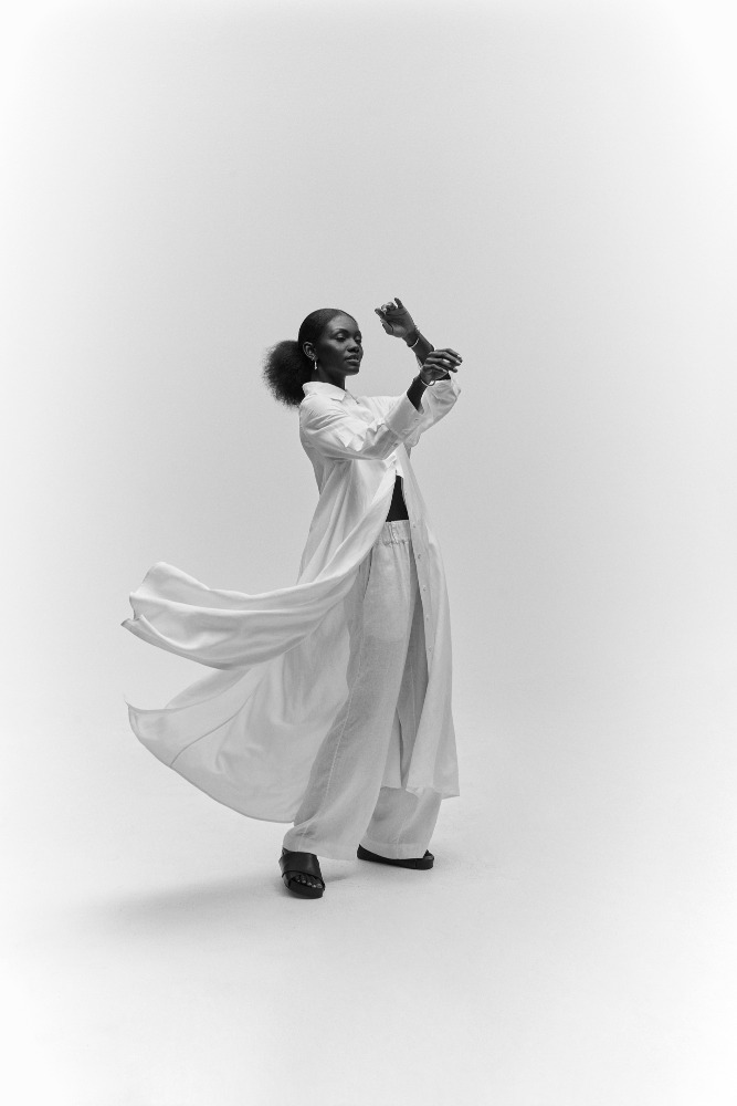 Elegant black female model in white, flowing shirt and palazzo pants strikes a dynamic pose against a stark white backdrop.