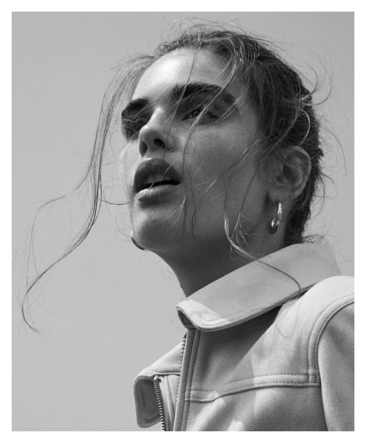 Monochrome portrait of a young woman in a light-colored jacket against a bright sky, looking upwards with her hair slightly disheveled.