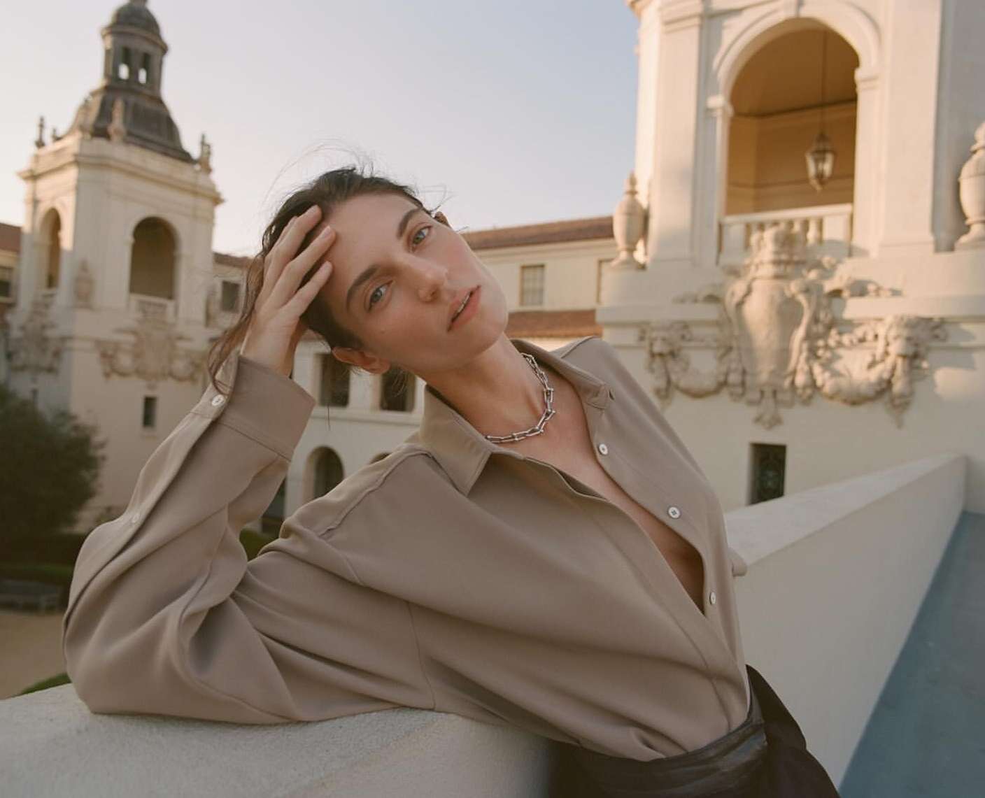 A woman in a tan shirt and silver necklace poses outdoors against a backdrop of a light-stone building with architectural details.