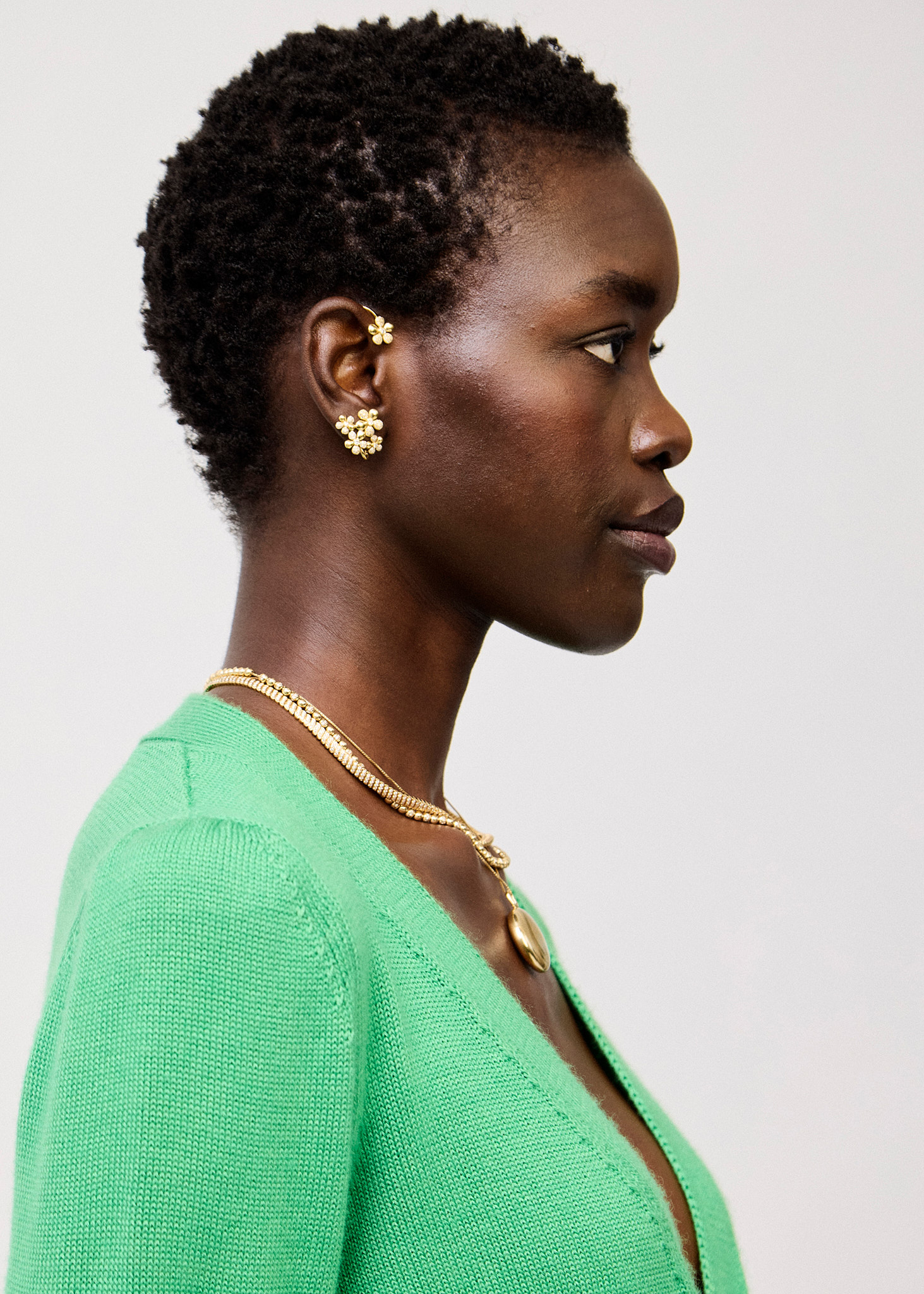 Profile shot of a Black woman with short natural hair wearing gold flower earrings and necklace, a green cardigan, against white.
