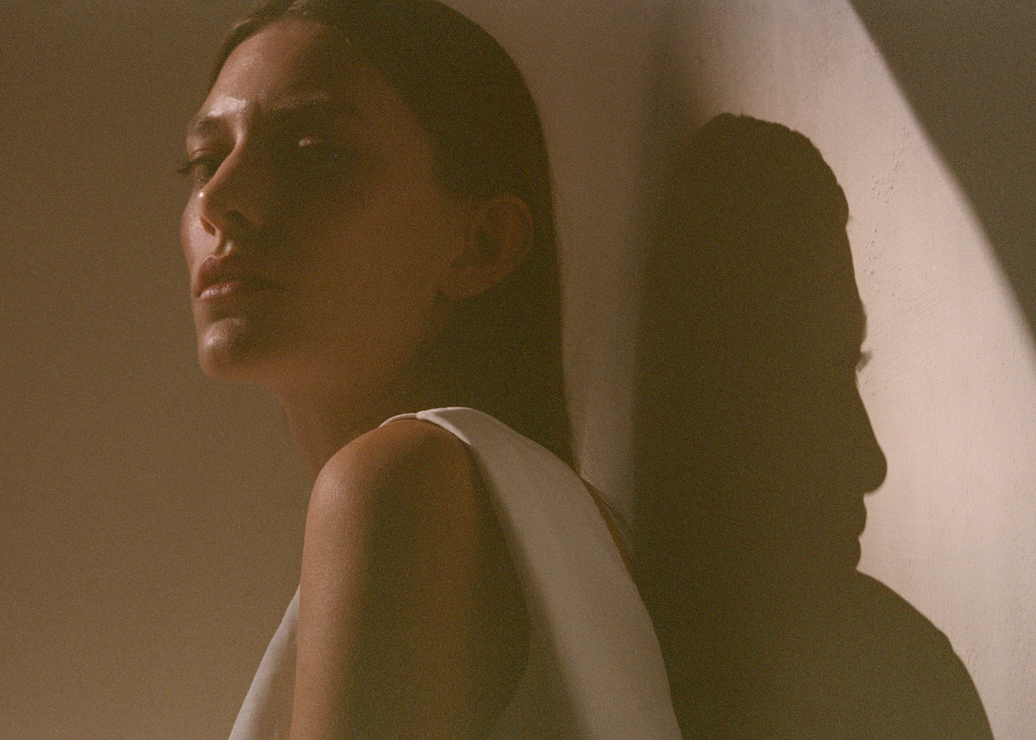 Portrait of a woman with long brown hair, wearing a white top, with a shadow on the wall behind her.