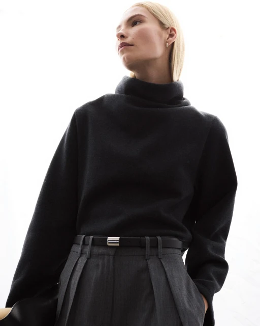 Blonde-haired woman in a high-neck sweatshirt and pleated slacks is pictured in a high-angle, waist shot against a bright background.