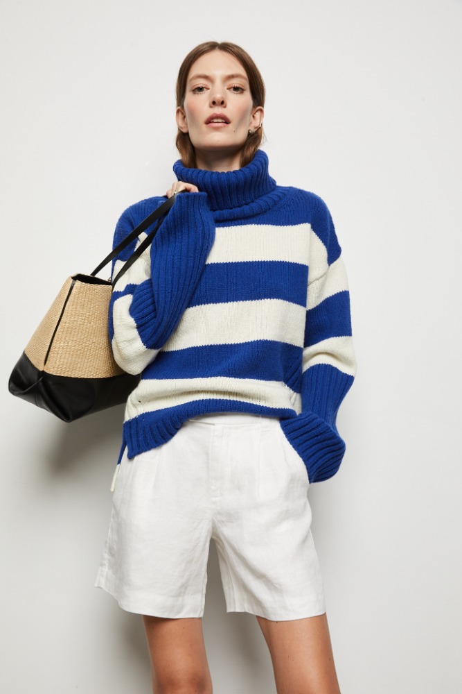 A woman models a blue and white striped turtleneck sweater and white shorts, holding a black and tan tote bag against a white backdrop.