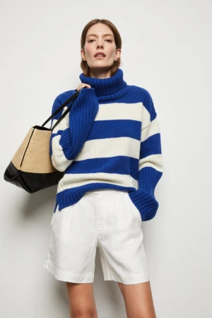A woman models a blue and white striped turtleneck sweater and white shorts, holding a black and tan tote bag against a white backdrop.