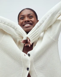Black woman wearing a textured white cardigan with arms raised laughing against a plain light background
