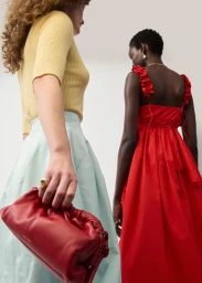 Two models posing against a plain wall, one wearing a yellow ribbed top and light blue skirt holding a red leather pouch bag.
