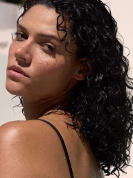 Close-up portrait of a woman with wet dark curly hair, freckles, and a black strap top against a bright background.