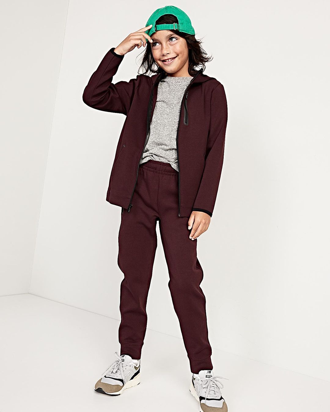 Child model poses indoors wearing a burgundy jacket and pants with a green cap, smiling, fashionably dressed against a white background.