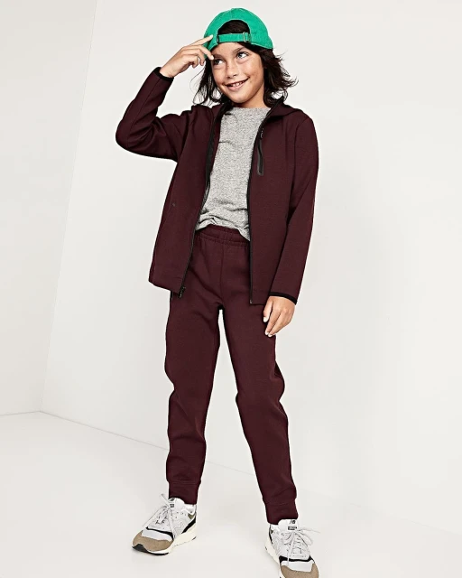 Child model poses indoors wearing a burgundy jacket and pants with a green cap, smiling, fashionably dressed against a white background.