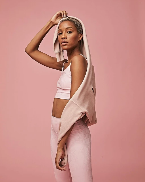 Woman with short hair wearing a pink athletic top and leggings and putting on a pink sweatshirt hood against a pink background.