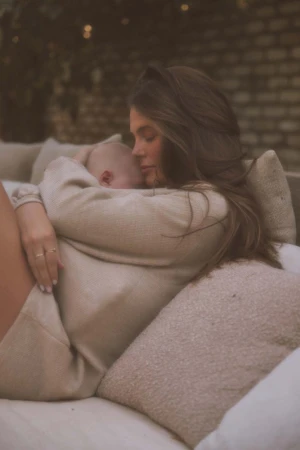 Woman in neutral knit sweater holding an infant while resting on outdoor cushions against a textured brick wall