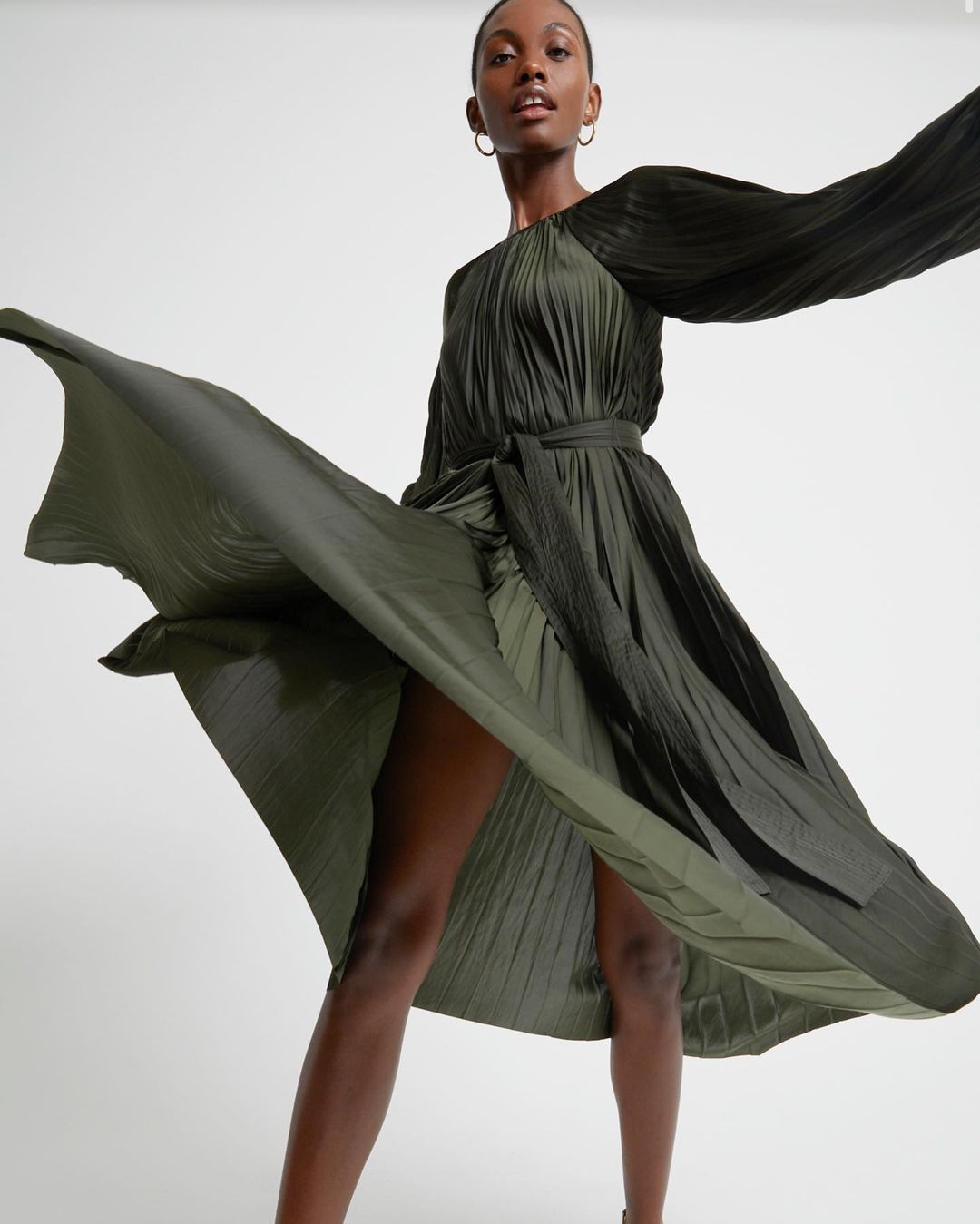 Elegant Black female model poses in a pleated olive-green dress, flared out, in a bright studio. Fashion, beauty, and editorial content.