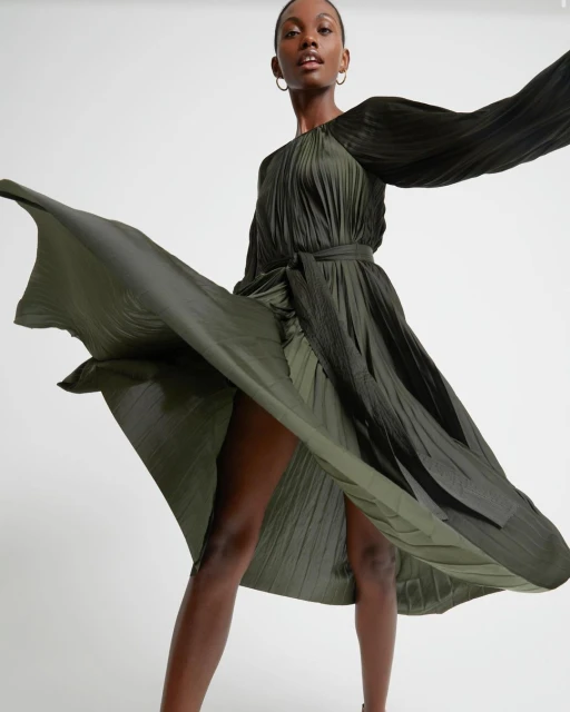Elegant Black female model poses in a pleated olive-green dress, flared out, in a bright studio. Fashion, beauty, and editorial content.
