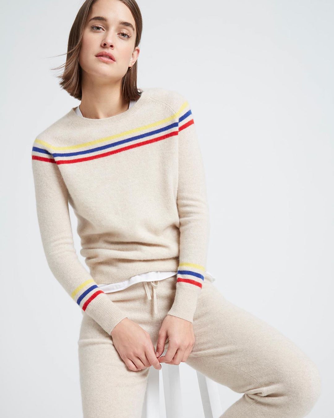 Woman in a beige sweater and pants sitting on a stool in a studio. Sweater has blue, red, and yellow stripes. Fashion shot.