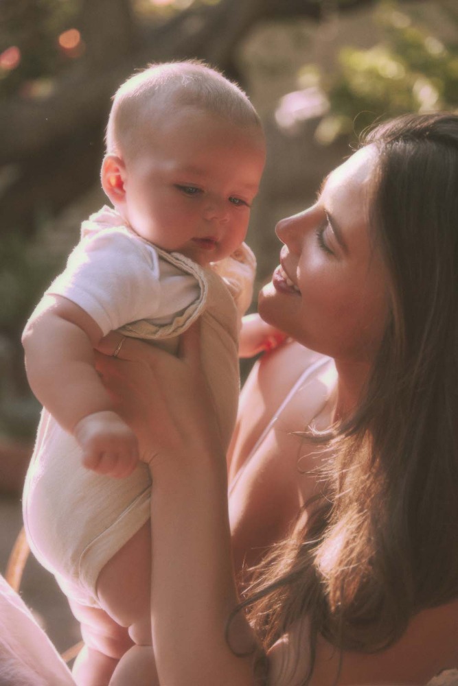 Portrait of a mother holding her baby outdoors in soft sunlight. The mother looks lovingly at the infant, who is held at chest height.