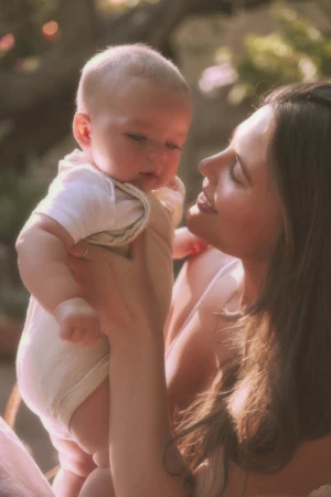 Portrait of a mother holding her baby outdoors in soft sunlight. The mother looks lovingly at the infant, who is held at chest height.