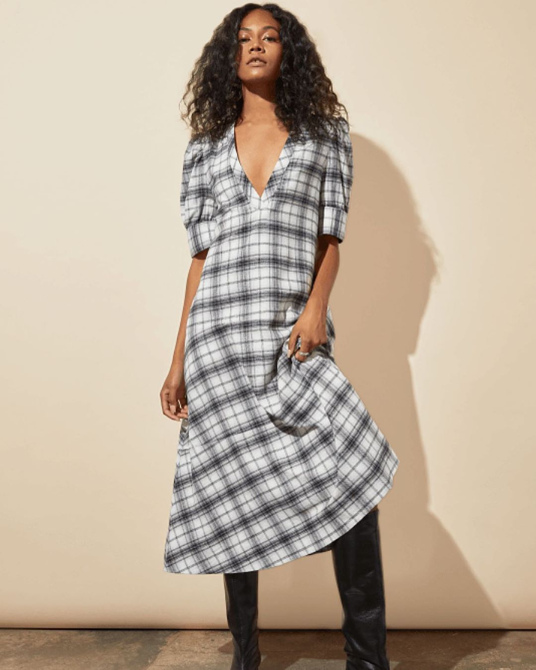 A young woman with curly hair poses in a flowing, plaid midi dress and black boots against a neutral backdrop in a fashion studio shot.