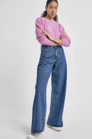 Woman wearing a pink knit sweater and blue wide-leg jeans with her arms crossed against a plain white background