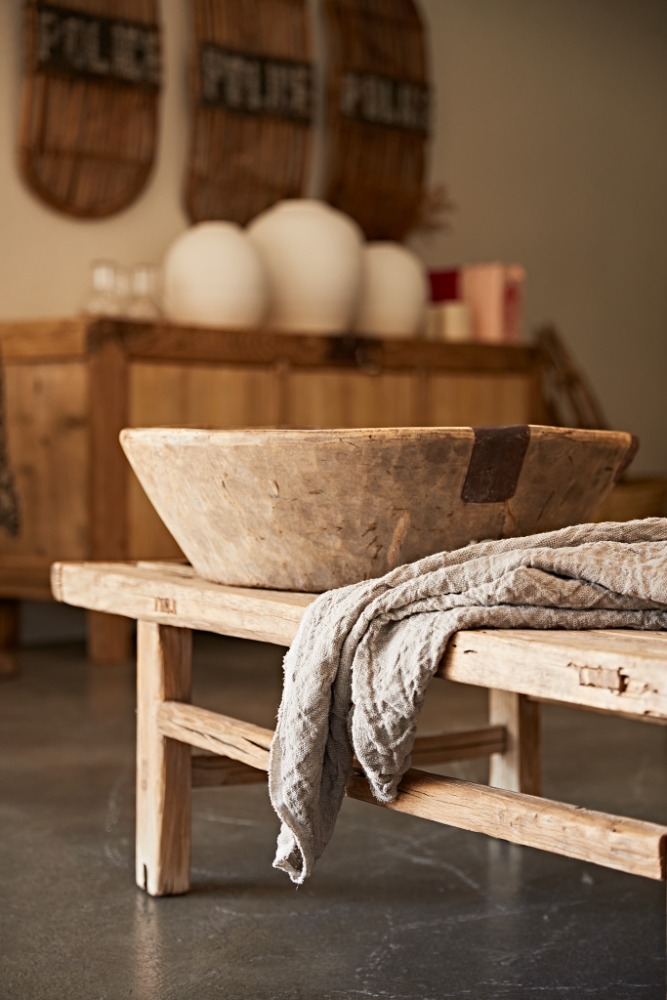 Rustic interior featuring a weathered wooden trough, a linen cloth, and vintage decor on a wooden bench against a neutral backdrop.