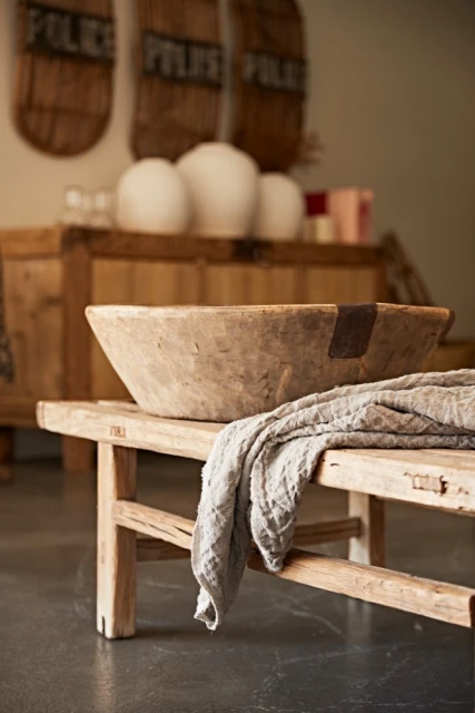 Rustic interior featuring a weathered wooden trough, a linen cloth, and vintage decor on a wooden bench against a neutral backdrop.