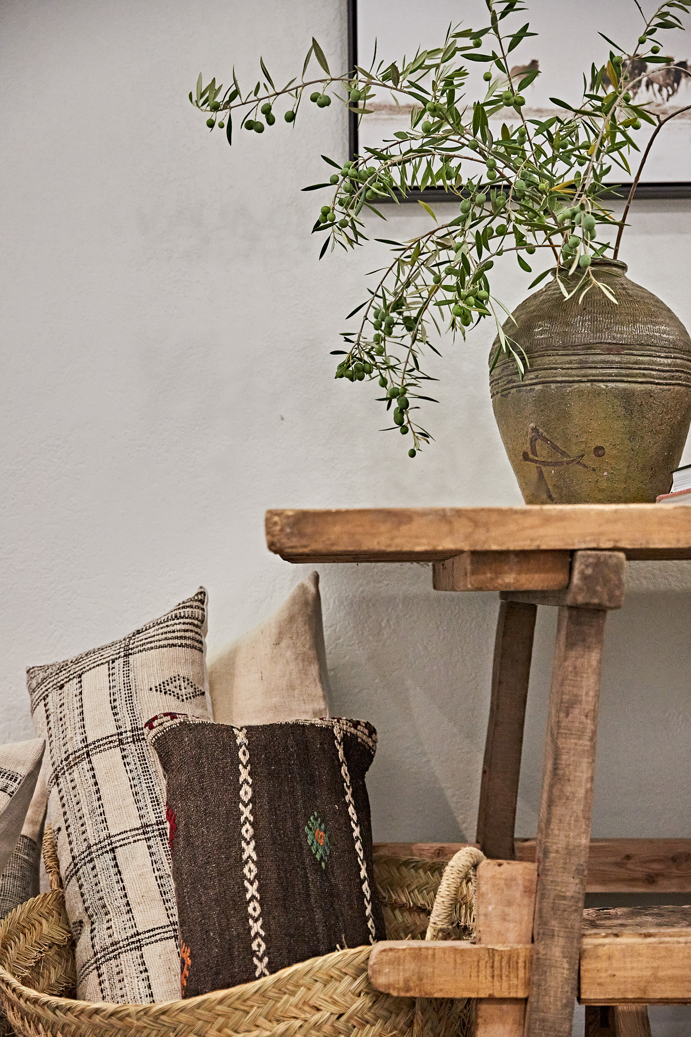 Still life of a rustic wooden table with olive branches in a vase, alongside woven baskets and decorative pillows against a neutral wall.
