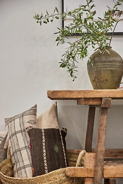 Still life of a rustic wooden table with olive branches in a vase, alongside woven baskets and decorative pillows against a neutral wall.