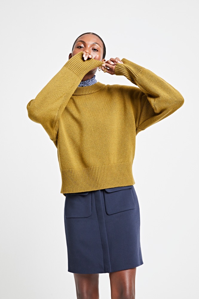 A woman in an olive sweater poses against a white backdrop, showcasing fashion apparel with a navy skirt, studio lighting, and style.