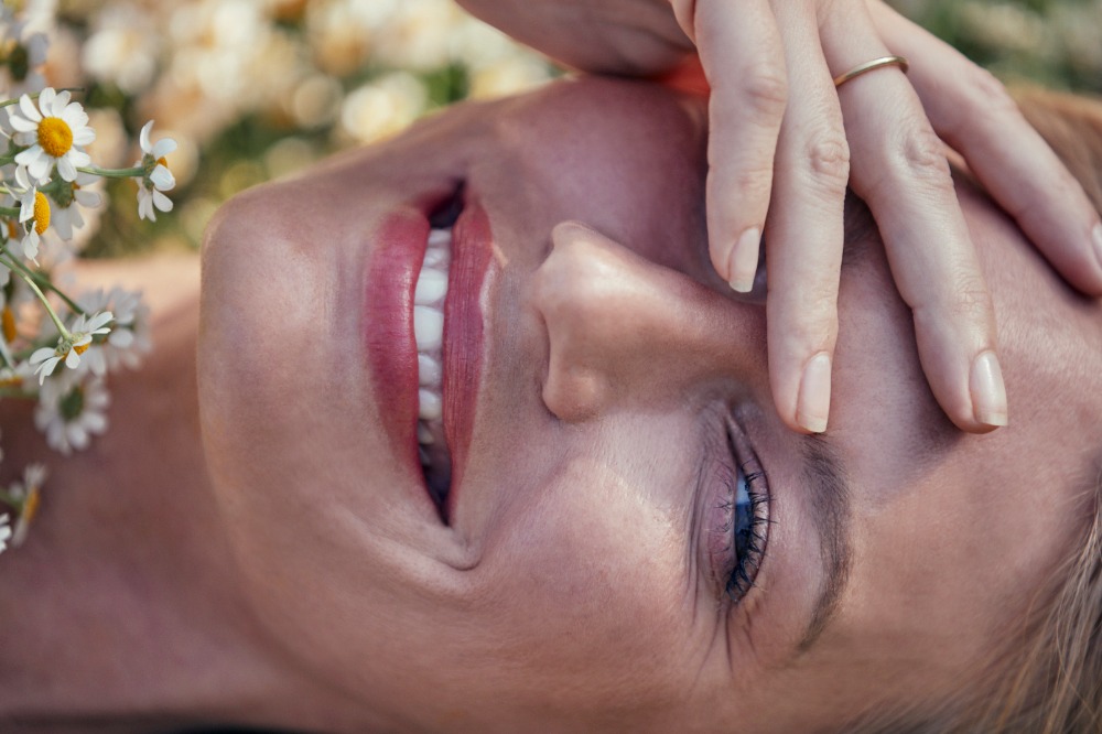 Close-up portrait of a smiling woman lying among wildflowers with her hand shielding her eyes