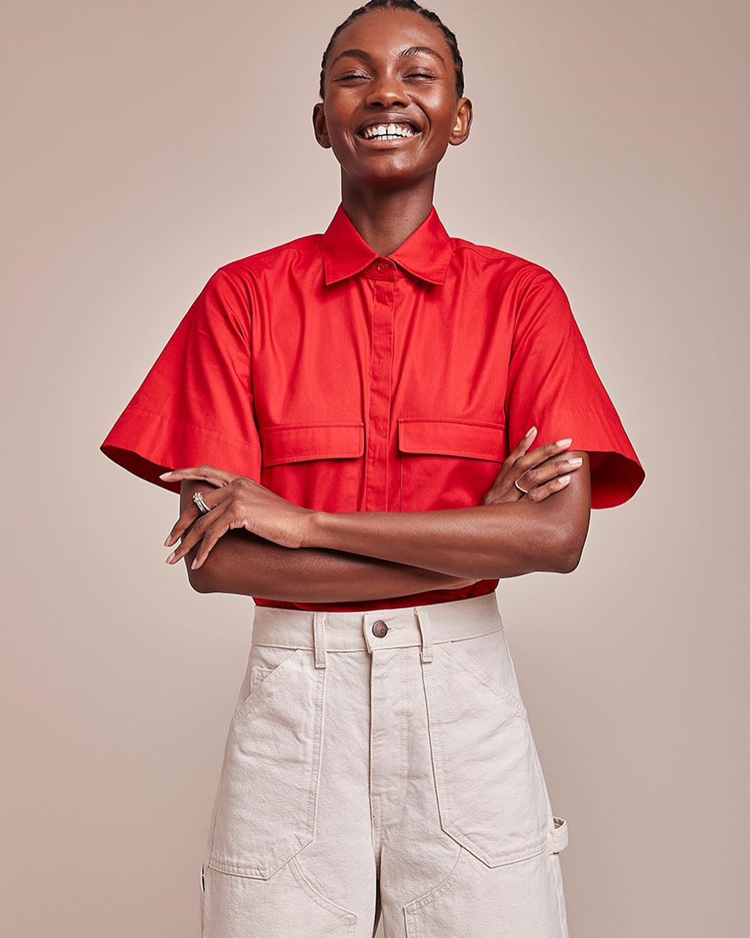 A smiling African-American woman in a red short-sleeved shirt poses in front of a neutral background, wearing beige carpenter pants.
