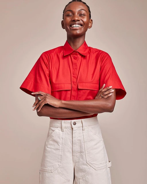 A smiling African-American woman in a red short-sleeved shirt poses in front of a neutral background, wearing beige carpenter pants.