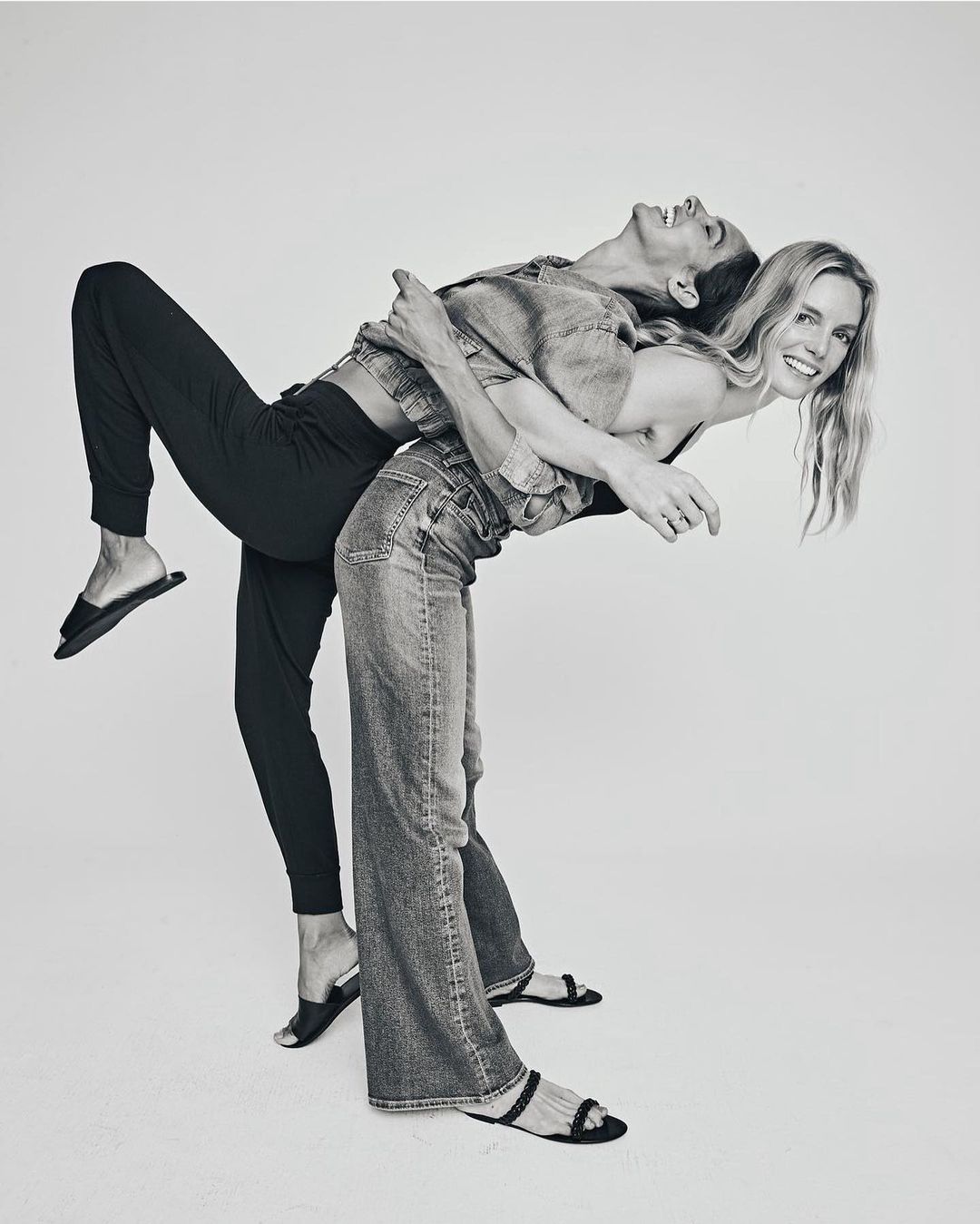 Two fair-skinned adult females are playfully posing back-to-back in a bright photo studio, showcasing casual denim and comfortable black pants.