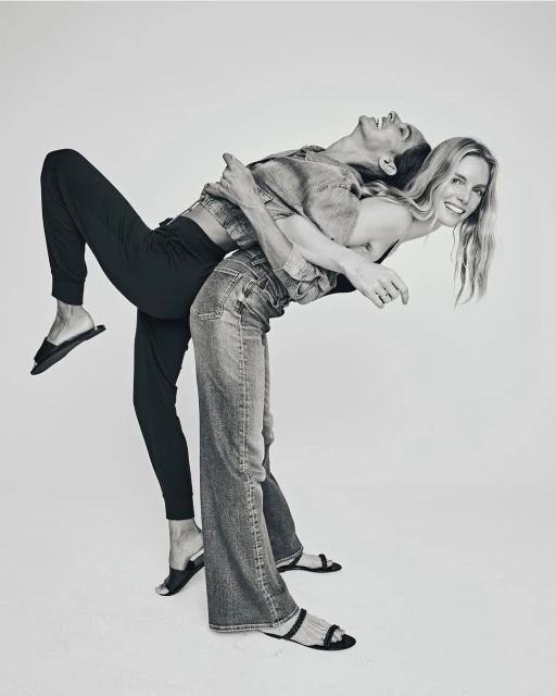 Two fair-skinned adult females are playfully posing back-to-back in a bright photo studio, showcasing casual denim and comfortable black pants.