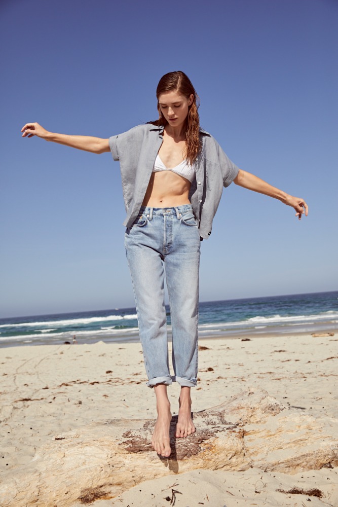Young woman in jeans and an open shirt stands barefoot on a beach. She has her arms outstretched toward the sky and ocean backdrop.