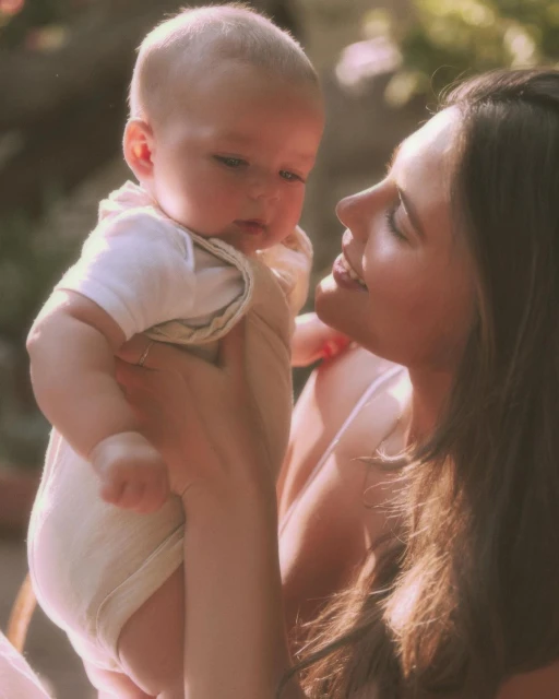 A mother gazes lovingly at her baby held in her arms in an outdoor setting with blurred green background, creating a heartwarming family portrait.