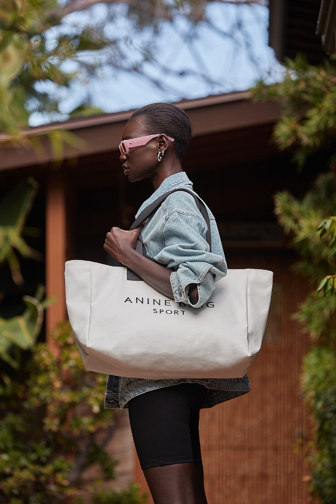 An adult female model is wearing a denim jacket and black shorts while holding a large tote bag in an outdoor setting with green foliage.