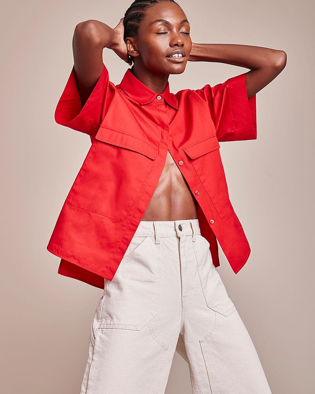 A dark-skinned woman poses indoors wearing a red short-sleeved shirt and beige trousers, gazing slightly upward with her hands behind her head.