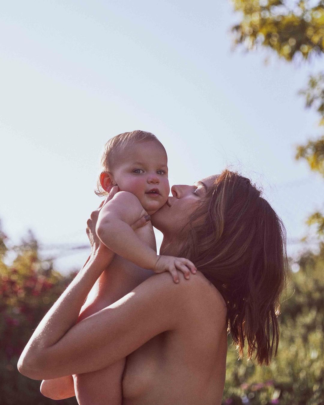 Woman kisses a baby on the cheek outdoors. The baby has a slight smile. The background features green foliage and a bright blue sky.