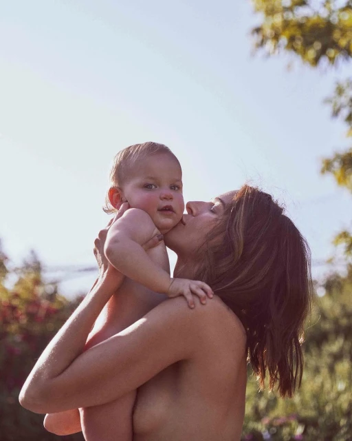 Woman kisses a baby on the cheek outdoors. The baby has a slight smile. The background features green foliage and a bright blue sky.