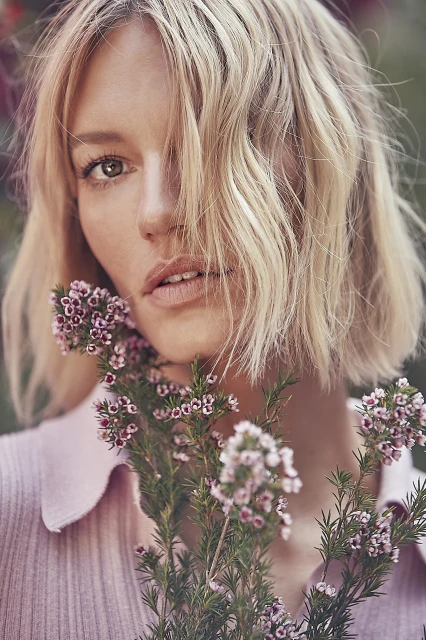 Close-up portrait of an adult female with short blonde hair, posing with pink flowers, wearing a pink collared shirt, and looking at the camera.
