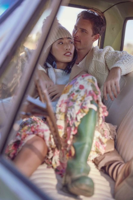 Couple in a vintage car with an Asian woman in a floral dress and green boots, and a Caucasian man wearing a white sweater.