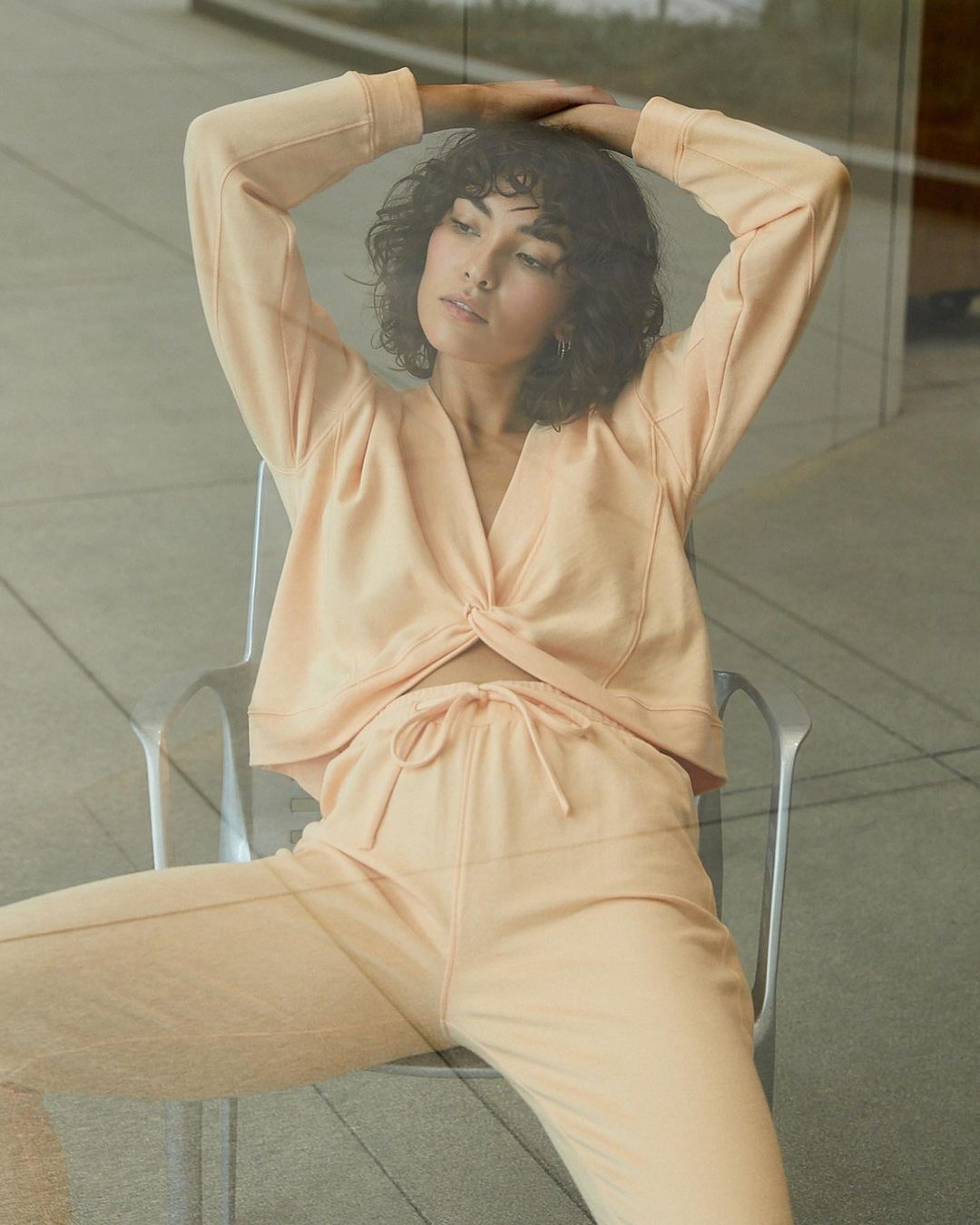 A woman with curly dark hair is sitting on a metal chair, wearing a matching peach-colored lounge set indoors with a blurred background.