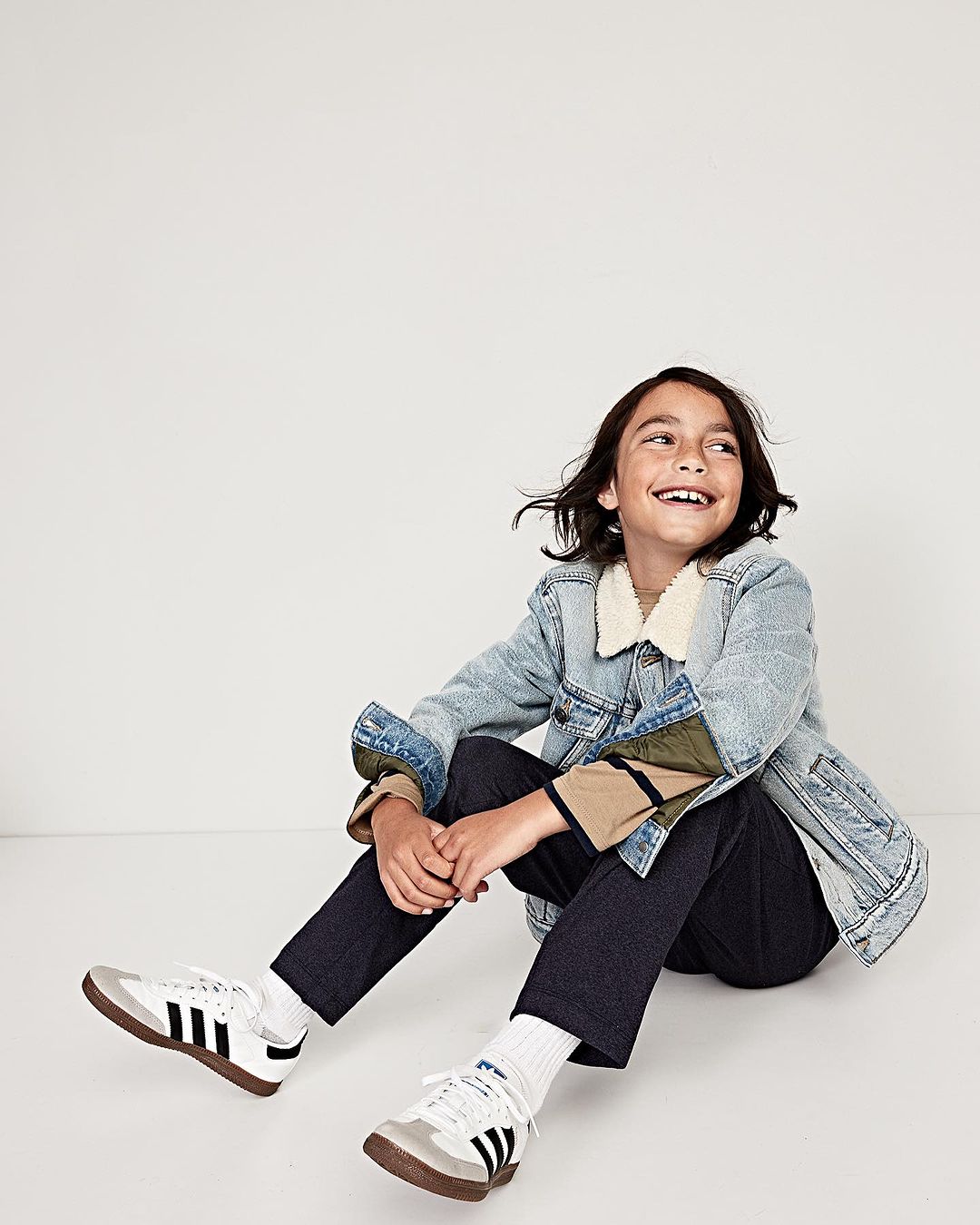 Smiling child sitting on a white studio floor wearing a denim jacket and black pants.